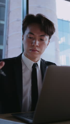 Asian businessman in formal suit works diligently on laptop marking notes on paper in modern office. Male financial specialist inspects data in professional atmosphere
