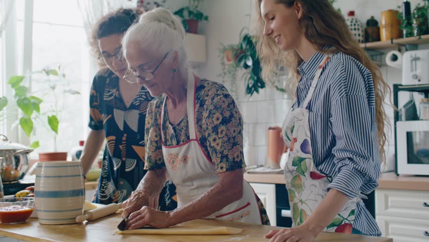 Elderly grandmother slicing pasta dough and talking as preparing dinner with family in cozy kitchen at home