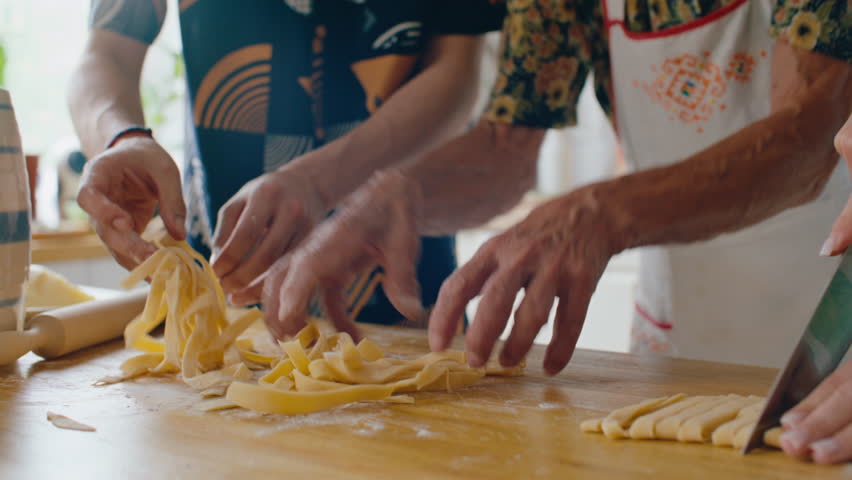 Hands of family members cutting, lifting and separating fresh pasta strips on floured wooden table while preparing homemade meal together in kitchen. Close-up shot