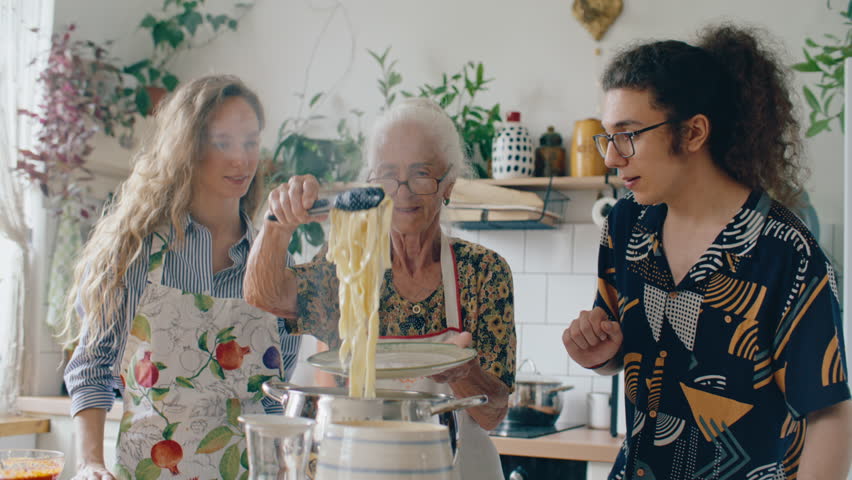 Elderly grandmother using kitchen tongs to serve freshly boiled pasta onto plate, smiling and chatting with two young grandchildren during family cooking time at home