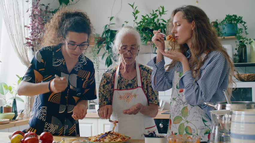 Senior grandmother and her young grandchildren tasting and enjoying freshly cooked homemade pasta, smiling and chatting in cozy kitchen