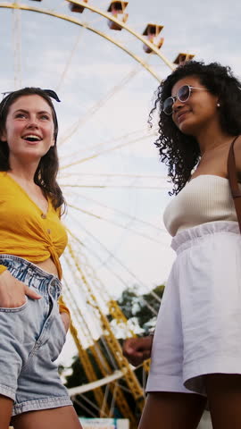 Two young alluring females in casual outfit are posing in park against a ferris wheel and blue sky. They are talking, smiling, seeing someone and waving hands. Summer day, green trees. Slow motion