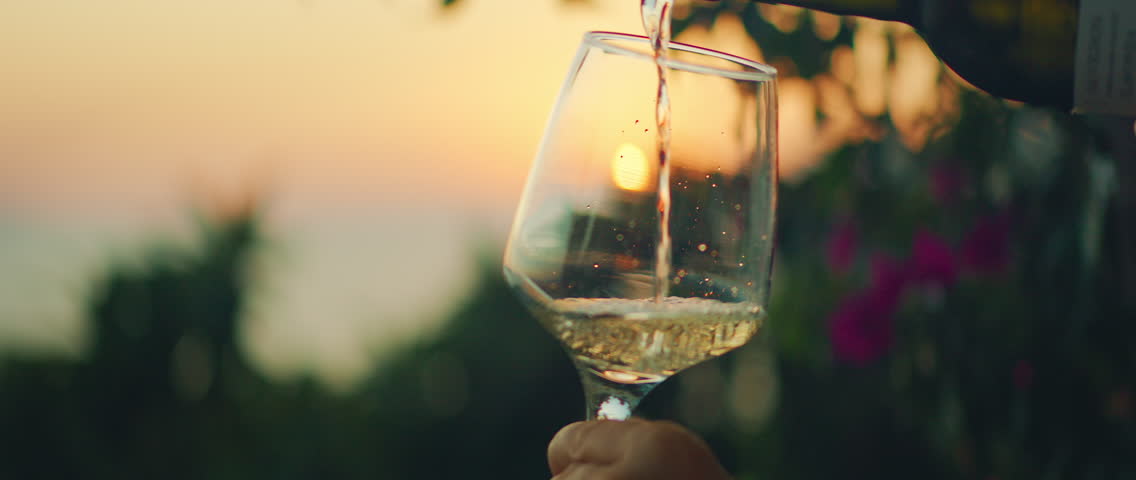 Close-up of white wine being poured into glass at sunset with sea and garden on the background