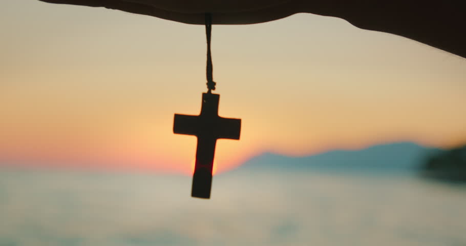 Hand holds wooden cross in silhouette against soft evening sky by ocean