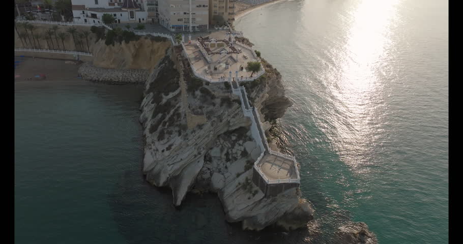 Drone footage shows the famous Balcón del Mediterráneo in Benidorm, Spain, perched dramatically on a white cliff above the turquoise sea. The viewpoint offers breathtaking coastal scenery.