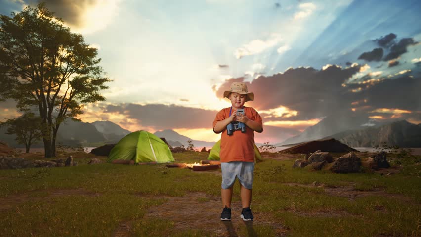 Asian Boy With A Hat And Binoculars Looking At Smartphone Then Smiling And Pointing To Side While Tent Camp Lakeside. Boy Researcher Examines Something, Travel Adventure, Full Body
