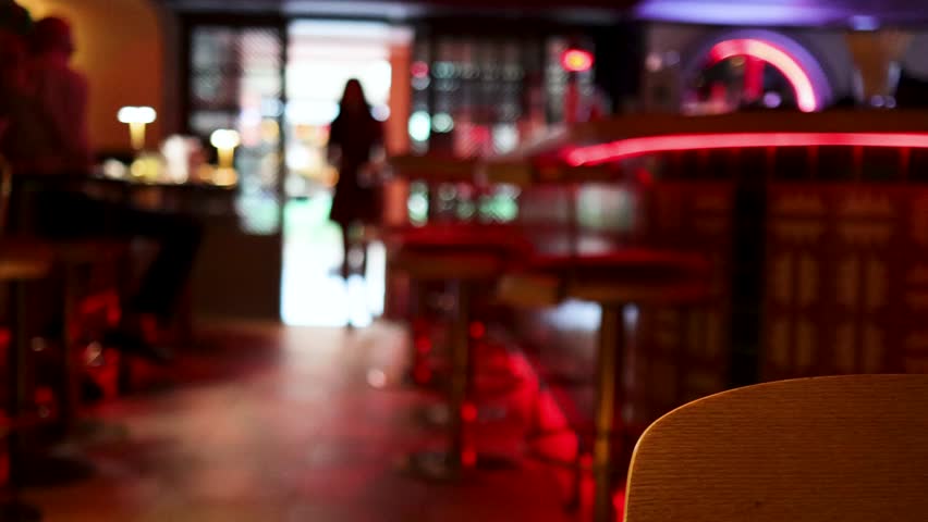 A silhouetted woman enters a contemporary bar with red neon accents, captured in soft focus with moody, low ambient lighting and a stationary camera