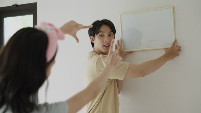 happy Asian couple standing together next to blank white wall of their new house, and wife trying to leveling framed picture while husband is hanging up with loving emotion.