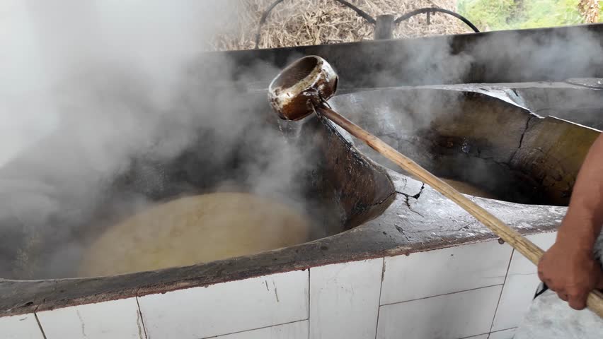 Steam rises from boiling sugar cane juice as a man scoops it from a heated vat