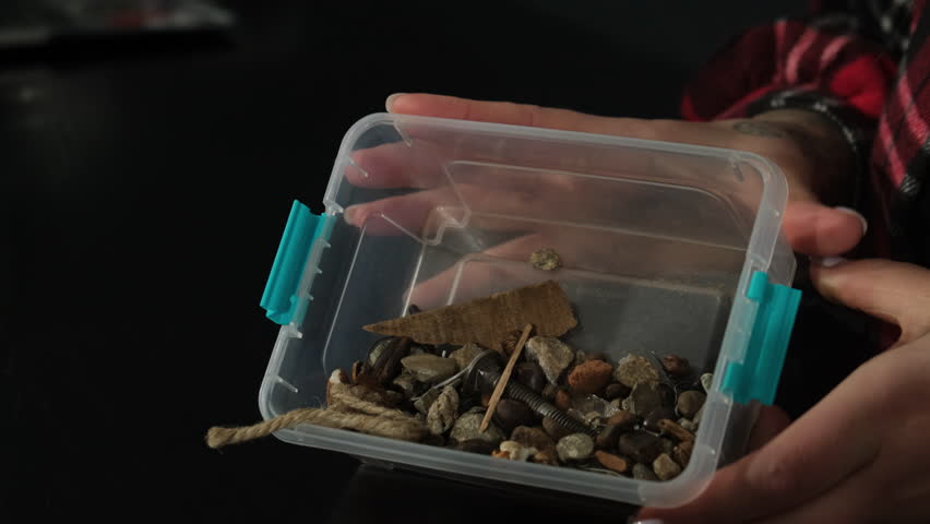 Woman holds clear plastic container filled with bric-a-bracs and gemstones. Lady shakes box in hands slowly pouring things onto table