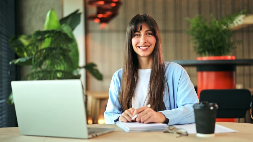 Cheerful professional businesswoman, dressed in a light blue shirt, works at a modern office desk. Focused on tasks, successful female smiles, feeling productive and pleased.