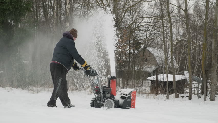 Man with gasoline snow blower clears road on village street.