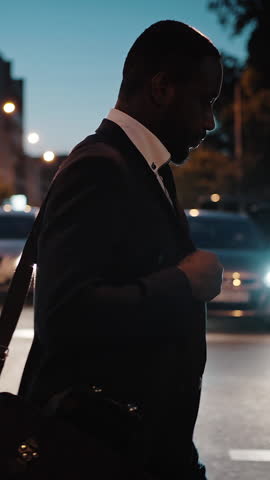 Night lifestyle. Tired african american businessman with bag passing road against cars, going home after hard working day, side view, slow motion