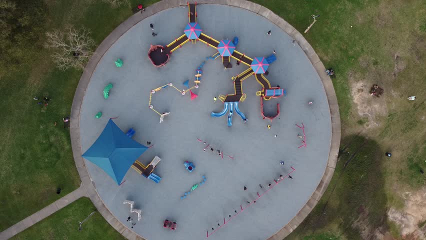 Kids swing, climb, and slide on a rounded concrete playground pad while parents supervise. Bright equipment and green lawns surround this lively outdoor scene in Clear Lake Park, Seabrook, Texas. USA