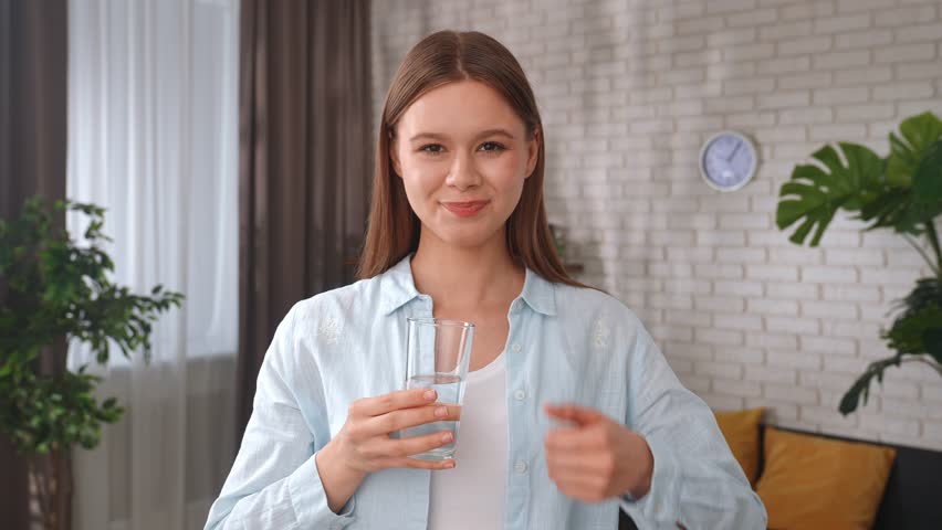 A young woman drinks a glass of fresh water indoors and shows a thumbs up gesture, emphasizing the importance of hydration and a healthy lifestyle.