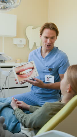 Vertical shot of smiling dentist explaining teeth whitening and holding tablet with pictures on screen