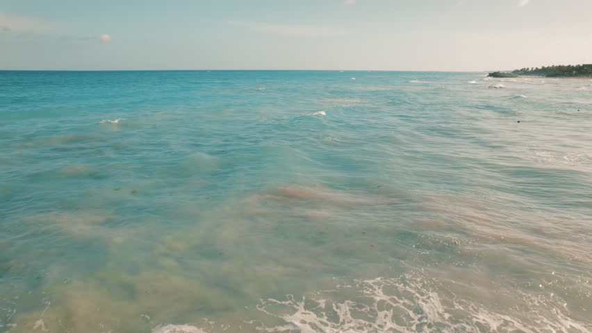 Woman in black dress stands alone on rocky point looking out over calm ocean small waves and sandy seabed in Tulum, Mexico