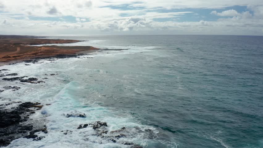 Aerial footage of Hawaii’s wild coastline reveals turquoise surf rolling onto red volcanic cliffs beneath a bright, cloud-streaked sky—capturing the island’s raw natural beauty and isolation.