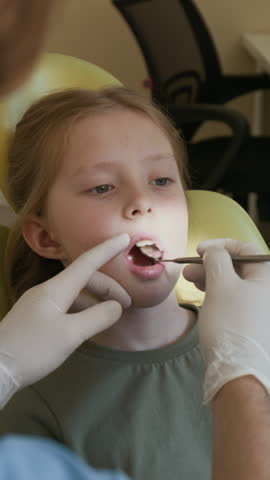 Vertical shot of cute young child sitting in dental chair while dentist using mirror to perform dental checkup