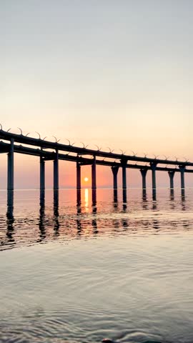 Beautiful sunset over the sea. The sun setting between the pillars of the bridge. Reflection of sunlight on water. Kanonersky Island, Saint-Petersburg. Vertical FullHD slow motion footage.