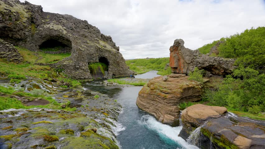 Stunning Gjain Canyon in Iceland spring featuring Eyeglass Cave and cascading water amid green mossy cliffs under a cloudy sky.