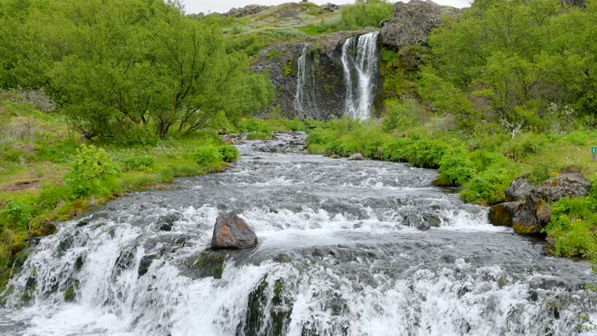 Vibrant Gjain Canyon waterfall flows through green valley with rocky cliffs under a cloudy sky in Iceland spring.
