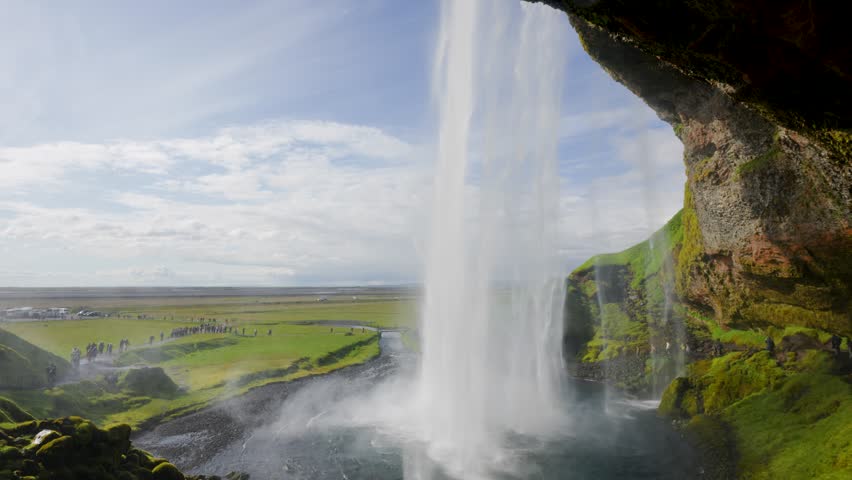 A unique perspective from behind Seljalandsfoss waterfall in Iceland, showing the powerful water curtain and misty spray with green cliffs and bright sky beyond.