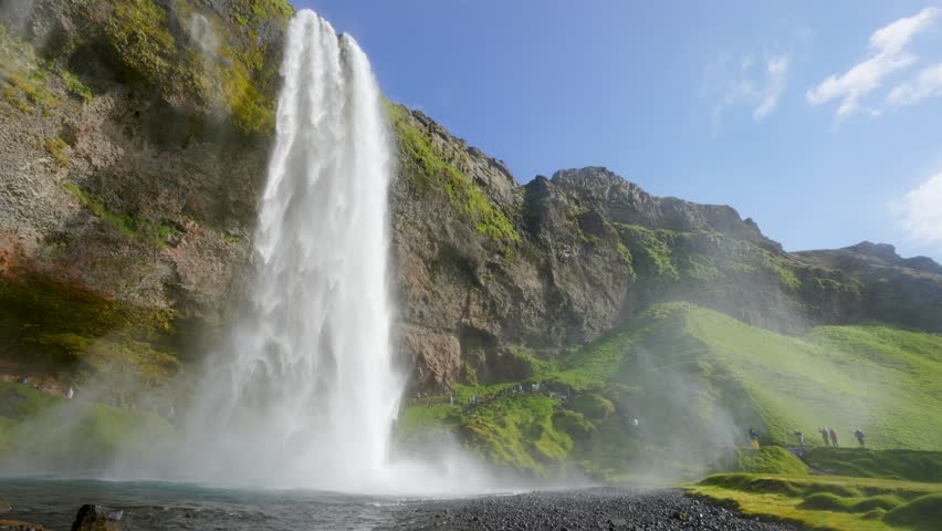 The iconic Seljalandsfoss waterfall in Iceland, featuring a vibrant rainbow at its base and surrounded by lush green cliffs under a clear blue sky.
