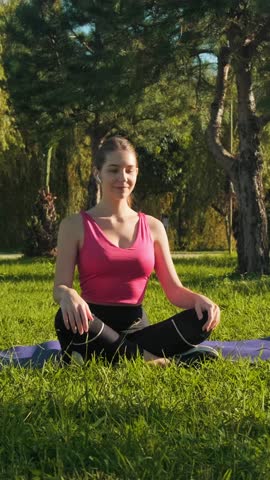 Young sexy woman in sportswear sitting on sports mat on the grass in the park and doing warm-up before main exercise or yoga. Female athlete tilting head to the right and left to stretch neck muscles