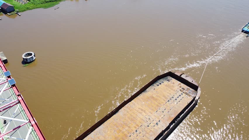 Aerial view of a tugboat pulling a barge filled with coal across the Rumpiang Bridge in the waters of South Kalimantan, Barito River