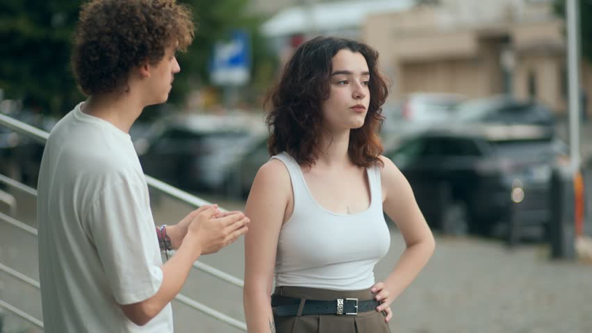 A young couple argues emotionally on the street. The man tries to explain and apologize as the woman walks away, visibly upset and unwilling to listen.
