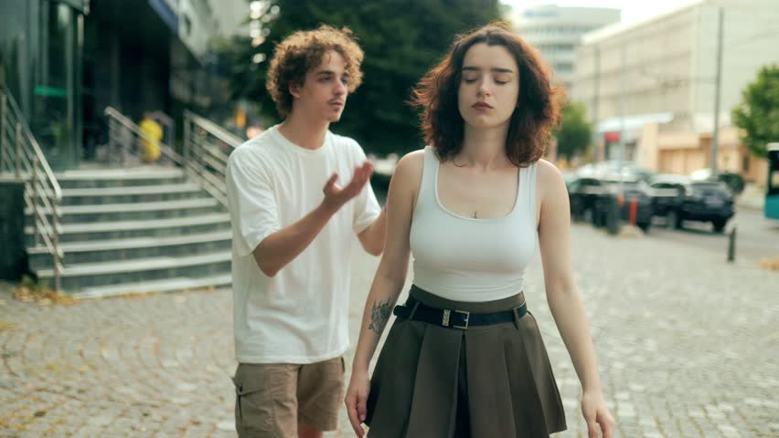 A curly-haired man kneels on the street, pleading emotionally with his partner. She stands with hands on hips, looking frustrated. Real urban couple moment outdoors.
