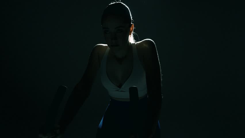Wide shot of a fit woman cycling in a dark gym, camera moving centrally toward her with a slight side slide as cinematic backlight highlights her shoulders and head, capturing power and rhythm