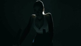 Wide shot of a fit woman cycling in a dark gym, camera moving centrally toward her with a slight side slide as cinematic backlight highlights her shoulders and head, capturing power and rhythm - Powered by Shutterstock - Get 15% off with code: PIKWIZARD15