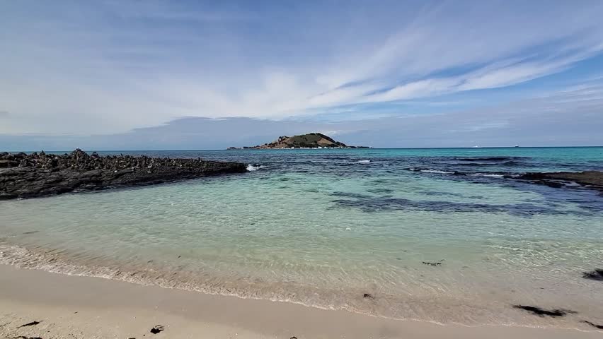 Clear emerald waves gently roll onto the white sand of Hyeopjae Beach in Jeju, Korea. Under a vivid blue sky, Biyangdo Island rises across the serene, transparent sea.