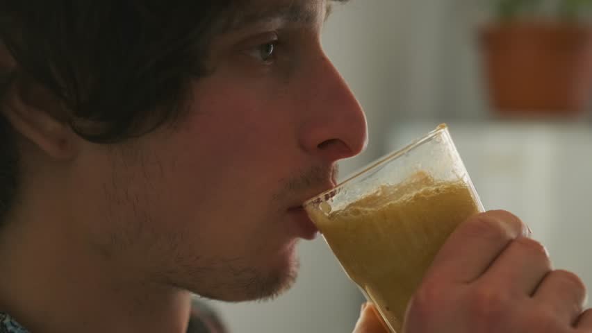 Side view of a young man drinking a thick homemade smoothie from a glass, enjoying a peaceful start to the day. Natural light and soft focus create a cozy, relaxed home atmosphere.