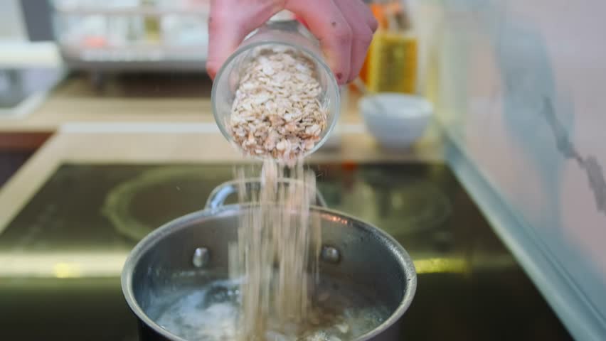 Close-up of male hand pouring raw rolled oats from a glass into a pot of boiling water on an induction stove. Start of a healthy breakfast cooking process in a minimalist home kitchen.