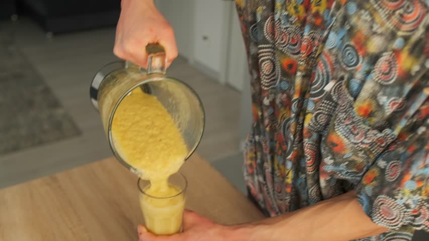 Close-up of a man in a colorful shirt pouring a thick yellow smoothie from a blender jar into drinking glasses in a bright modern kitchen. Healthy homemade drink preparation with natural ingredients. 