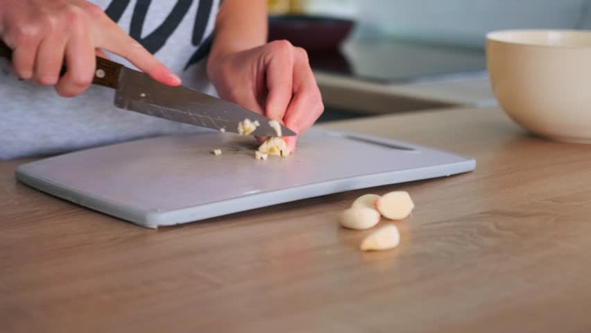 Woman hands chopping raw garlic on a scratched plastic cutting board with a large kitchen knife. Cooking with fresh ingredients at home.