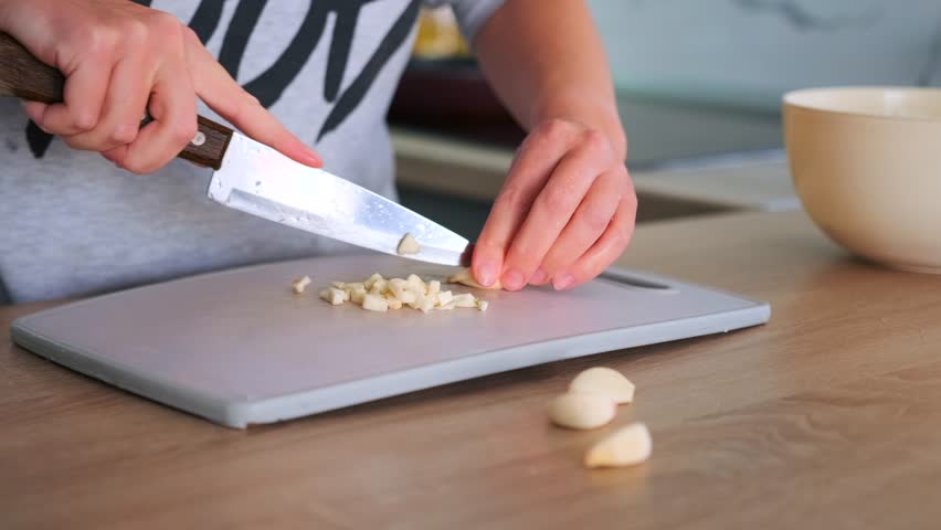 Woman hands chopping raw garlic on a scratched plastic cutting board with a large kitchen knife. Cooking with fresh ingredients at home.