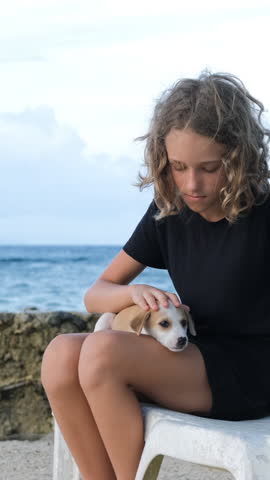 Vertical video. Young girl sits on a sunbed, cradling a cute puppy on her knees and gently petting the dog, relishing the serene coastal atmosphere, slow motion