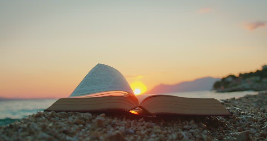 Book pages flutter on rocky beach at dusk with the sun setting behind mountains