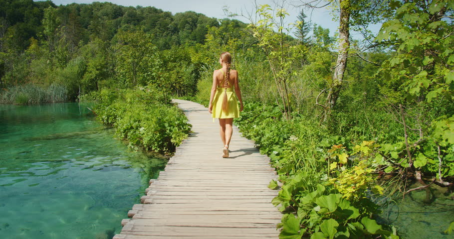 Woman walks alone along wooden path beside turquoise lake in summer Plitvice Park