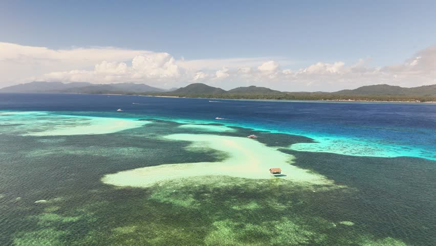 Drone captures Candaraman Sand Bar in Sulu Sea with rustic nipa-style floating hut perched over shallow coral waters, backed by white sandbanks and dense tropical forest, Balabac Island, Philippines.