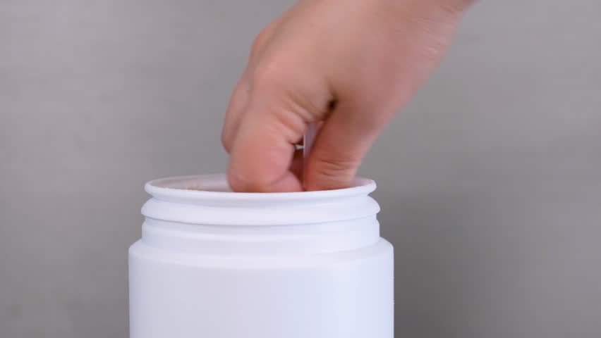 Hand takes protein powder in scoop from plastic jar on gray background close-up.