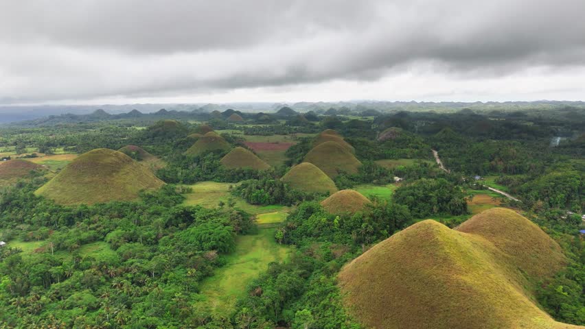 Undulating terrain of the Chocolate Hills in Bohol, Philippines with lush vegetation and cloudy skies, featuring symmetrical grass-covered mounds dotting tropical countryside stretching to the horizon
