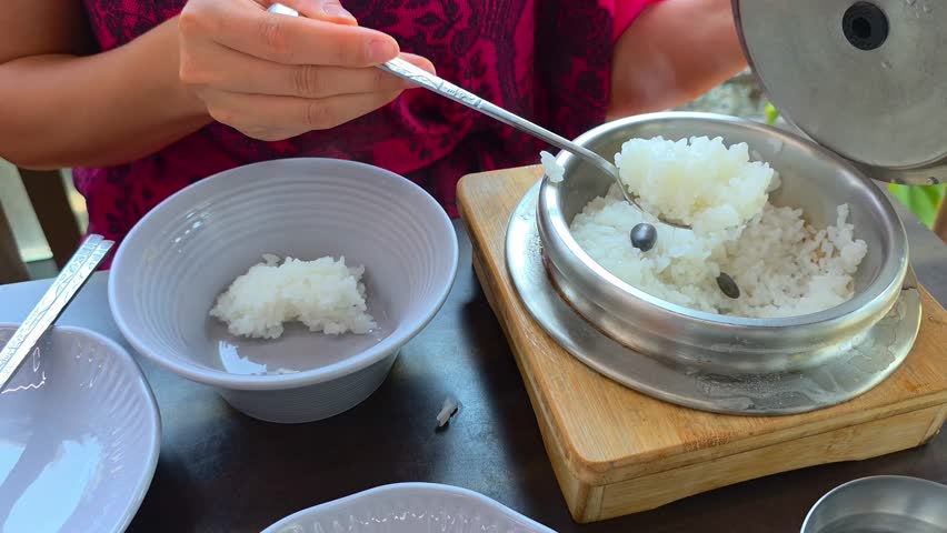 A close-up of a Korean woman traditionally eating rice by scooping hot rice from a metal bowl and pouring hot water to enjoy the remaining scorched rice.