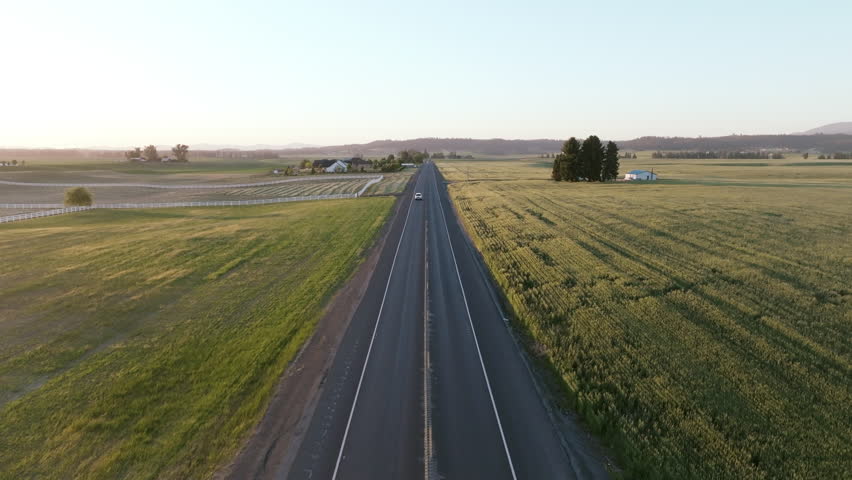 A lone car travels down a rural road dividing golden farmland and homesteads in the warm light of evening.