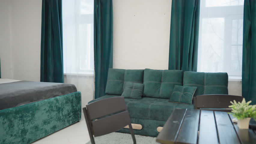 Modern bedroom with green velvet bed frame, plush gray bedding, teal curtains, and cozy green velvet couch with cushions. Bright natural light coming through large window