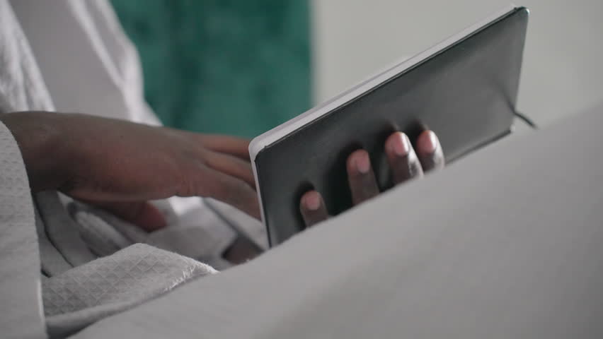 Close-up of man in white bathrobe holding black notebook, turning pages, soft light illuminating hands, green velvet headboard in background, relaxed indoor setting